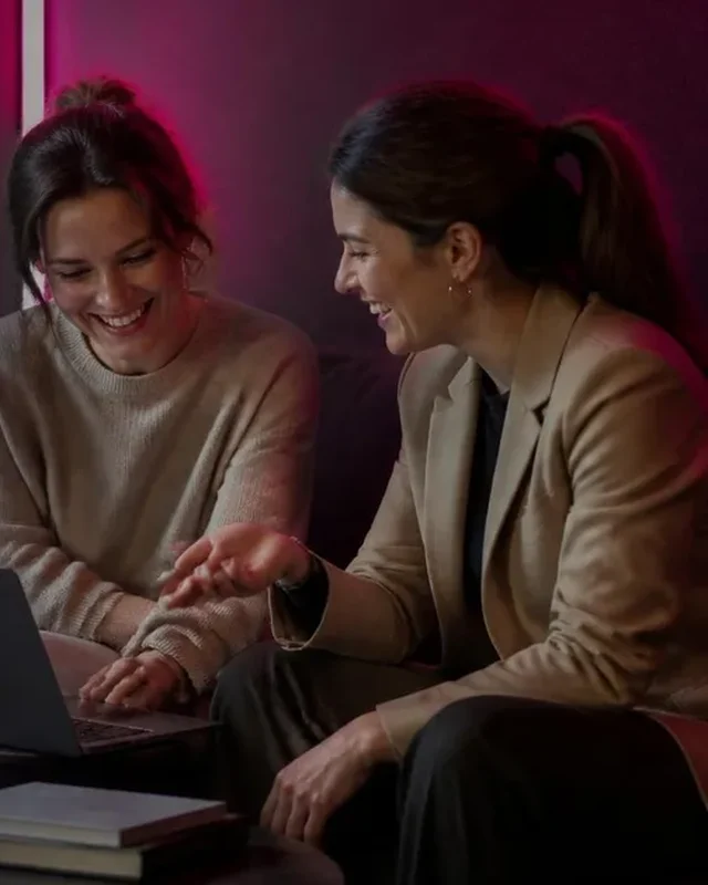500 Real Fans Beat 50,000 Lurkers: Girls Lounge cover image showing two women brainstorming together over a laptop on a sofa, neon pink lighting (AI-generated illustration)