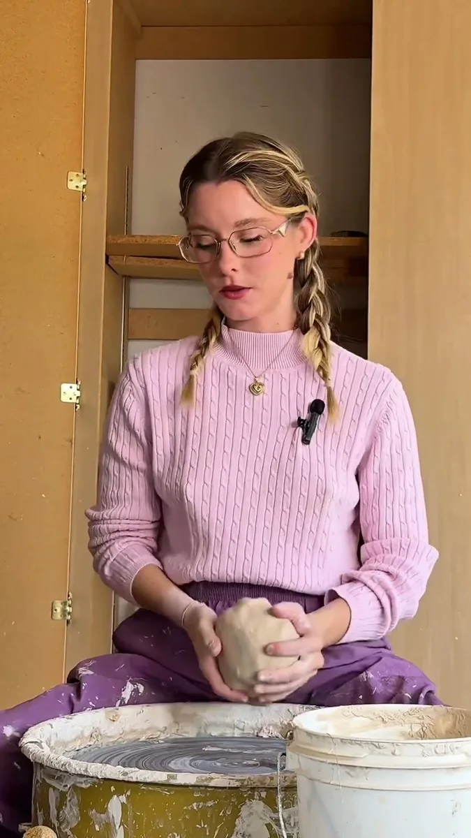 Haven Tunin in a pink sweater and glasses, holding clay at her pottery wheel